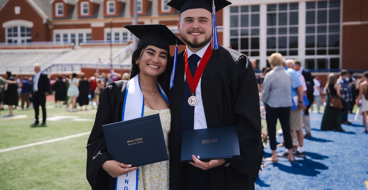 Female and male student smiling and posing with their diplomas on football field